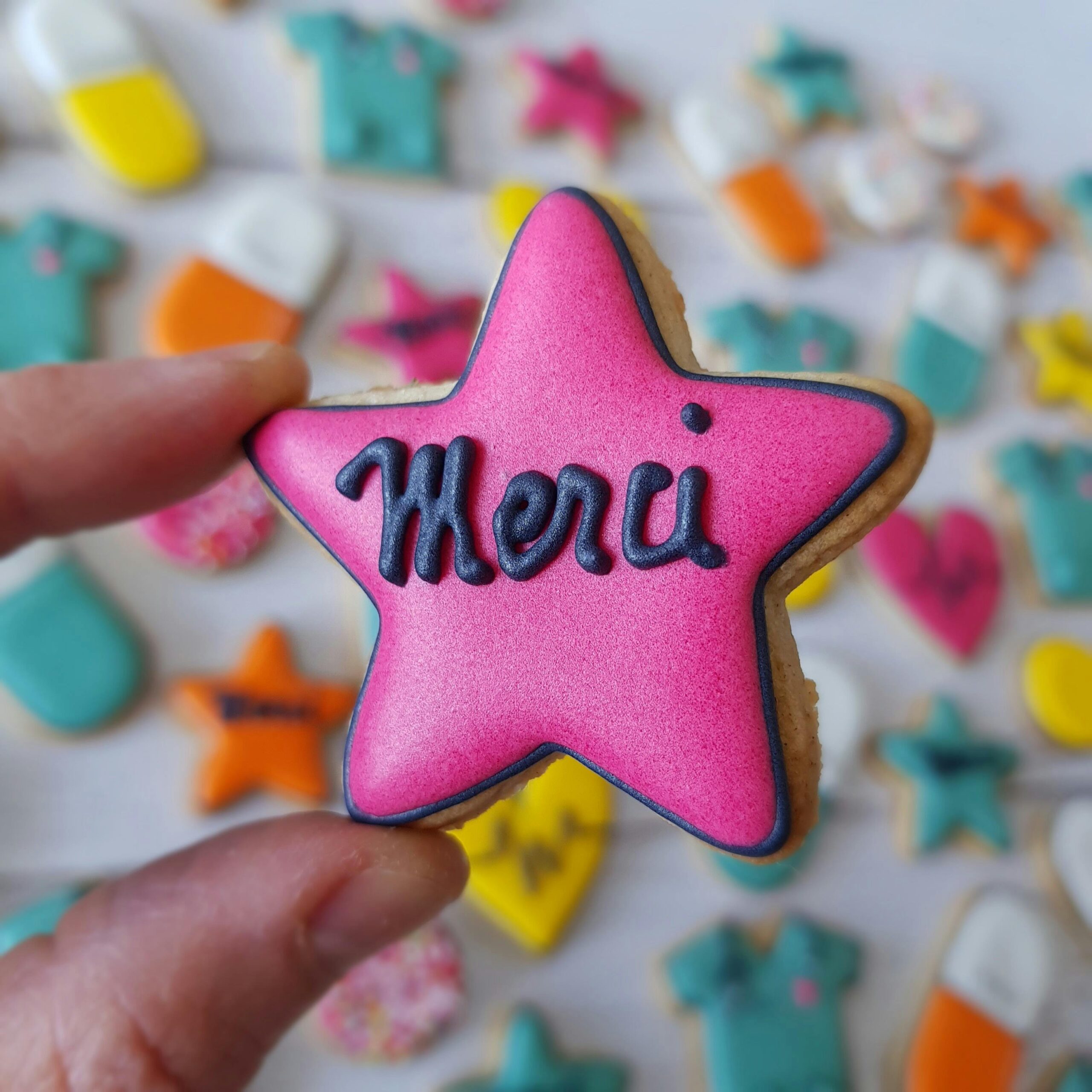Close-up of a pink star-shaped cookie with 'Merci' icing amidst a variety of colorful cookies.
